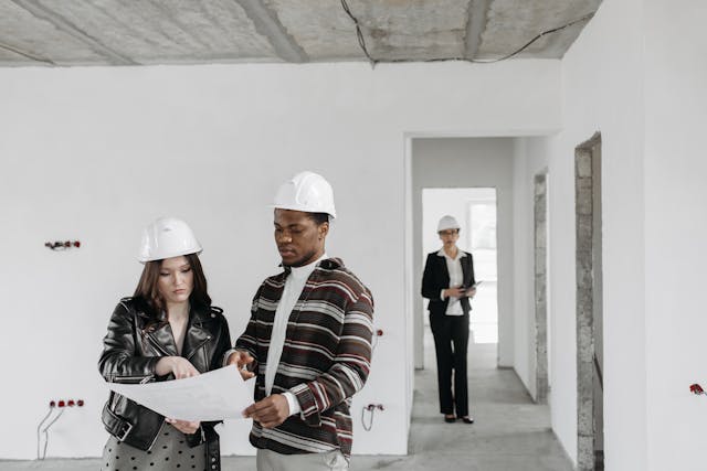 two people in white hard hats looking over a floor plan in an empty white house while a third person wearing a white hard hat looks on